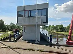 Brughuisje in parallellogramvorm met de kleuren wit, rood (deur) en bruin (raamkozijnen) (mei 2022)