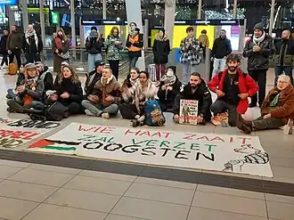 Kleine sit-in op Utrecht Centraal Station. Op de achtergrond zitten en staan pro-Palestijnse demonstranten, op de voorgrond ligt een spandoek met de tekst "Wie haat zaait zal verzet oogsten" en een kleine getekende Palestijnse vlag. Daarachter een kleine poster met de tekst "Resistance is justified when people are occupied".