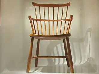 Foto van een houten stoel met een rugleuning met spijlen. Halverwege de rugleuning is een boog die overloopt in de armleuningen. De foto van de stoel is gemaakt tegen een lichtgekleurde achtergrond.