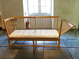 Foto van een ruimte in het Deens designmuseum in Kopenhagen met daarin een houten bank. De rugleuning en armleuningen van de bank hebben spijlen. De zitting heeft een gecapitonneerd kussen met een tattersall ruitpatroon. De rechter armleuning van de bank is door middel van een touw in een hoek van 45 graden gezet.