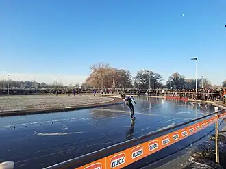 Schaatser op een buitenschaatsbaan. Publiek kijkt toe.