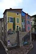 Trompe-l'oeil op de hoek van de Quai Vailhe-Planol en de Rue Droite. De muurschildering suggereert een huis op een fundament van natuursteen, met een man op een balkon.
