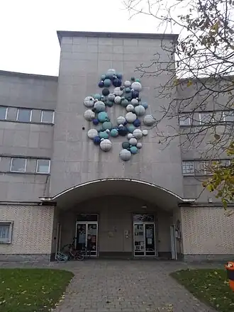 Centraal deel van gevel van een modern schoolgebouw. Boven een poort bevindt zich een kunstwerk met een wolk van blauwe en witte bollen.