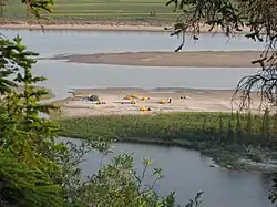 Canoes and tents rest on a sandy spit along a river.