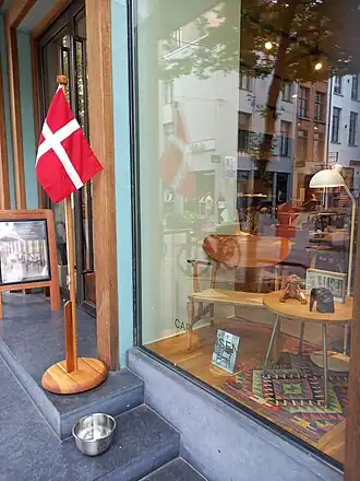 Foto van de etalage van de winkel van Carl Hansen & Søn. In de etalage zijn een aantal houten meubelen te zien. Naast de ingang van de winkel staat een Deense vlag.