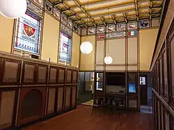 Interieur De Utrecht, 2023. Gebrandschilderde ramen met vestigingsplaatsen van (bij-)kantoren van De Utrecht, rijk versierde kasten aan weerszijden van de vloer.