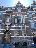 Pand in neorenaissance trant met trapgevel, waarin ontlastingsbogen, blokken, banden. Links een terugliggende ingangstravee met boogfries en balcon met balustrade