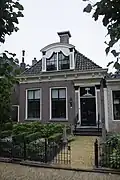 Dorpshuis met door pilasters geflankeerde ingang onder schilddak met grote versierde dakkapel en twee topschoorstenen met borden