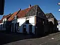 Hoekpand met gepleisterde lijstgevel, dakvenster met vleugelstukken. Stoeppalen met keitjesstoep