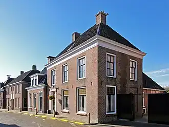 Breed pand met omlijste ingang, verdieping met zesruitsvensters en geprofileerde kroonlijst onder schilddak met hoekschoorstenen