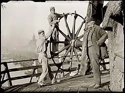 Molenaar Gerardus Hendrik de Boer en twee knechten bij de kruias van korenmolen De Gooyer. Gezien in noordelijke richting naar Nieuwevaart. Op de achtergrond de Oosterkerk; 1 november 1896, gefotografeerd door Jacob Olie.