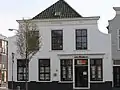Huis met gepleisterde ingezwenkte lijstgevel; hoge kap. Links winkelpui