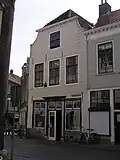Huis met gepleisterde gevel, verminkte top, en gedeeltelijk verminkte vensters. Winkelpui met omlijste middendeur. Kelderluik