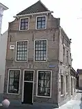 Hoekhuis met gesausde afgeknotte topgevel. Geprofileerde waterlijst en staafankers. Twee 24-ruitsvensters. Links een aanbouw. Zijgevel aan de Vlasmarkt met jaartalankers en gevelsteen voorstellende St. Sebastiaan