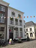 Huis op de hoek van de Hamstraat, met gepleisterde lijstgevel, waarin ingangspartij met fraai bovenlicht uit ca. 1800 en kuiven van stuc boven de segmentbogen der vensters. Nog geheel woonhuis
