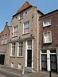Huis met witgeverfde gevel en steil, aan de voorgevel afgewolfd, zadeldak. Hoge kelder, schuiframen, deuromlijsting, lelie-ankers