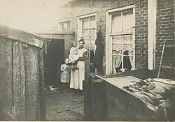 Moeder met kinderen voor haar woning in de Davidsteeg in 1914. Gemaakt in opdracht van de gemeente in het kader van het verbeteren van de hygiënische toestand in deze slop. Het bijschrift luidt 'De waterafvoer is gebrekkig; bij stortregens loopt het water de woningen in.'