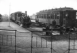 NS-rijtuig 3de klasse met links de aankomst van stoomlocomotief nr. 7003 van de NS (serie 7000) met een rijtuig en goederenwagen op het Station Amsterdam Haarlemmermeer.