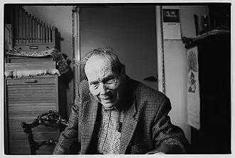 Foto van fotograaf Hans Sibbelee op ca. 86-jarige leeftijd. De oude man met sterk gegroefd gezicht zit aan een tafel in de voorkamer van zijn huis. Hij zit midden in beeld en kijkt vriendelijk lachend naar de camera. Zijn hoofd en bovenlijf zijn te zien. Hij draagt een ruimvallend colbert van een tweedstof en een geblokte bloes. Zijn bril steekt in zijn linkerborstzak. Achter de fotograaf is - van links naar rechts - een houten kast met roldeurte zien, er staat een houten stoeltje voor de kast en een houten orgeltje bovenop. Rechts ernaast zien we een deur en weer daarnaast de donkere kast van een wandpiano waar een aantal blokfluiten op staat. Tussen de deur en de piano hangt een kalender met honden aan de muur. Tegen de zijkant van de piano hangt een kalender van een chinees restaurant.