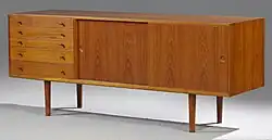 Foto van een teakhouten dressoir met vijf laden en twee schuifdeuren. De foto van het dressoir is genomen tegen een witte achtergrond.