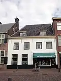 Woonhuis-winkel. Pand met wit blokpleisterwerk en gesmoorde oud-hollandsepannenkap met twee dakkapellen.