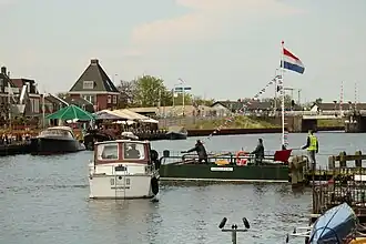 Het veer begint zonder passagiers aan zijn overvaart, terwijl de loods (met rode vlag) een schip tegenhoudt. Dit schip kan pas verder varen als de kabel neergelaten is. Op de achtergrond de oude brug in de Burgemeester Stramanweg (N255).