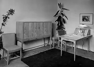 Zwart-wit foto van een meubelopstelling tijdens de tentoonstelling van het meubelmakersgilde in 1937. Op de foto worden meerdere houten stoelen, een houten dressior en een houten bureau getoond.