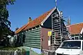 Dorpshuisje, onder zadeldak tussen houten topgevels; hoge schoorsteen. Houten zijwanden. Voorgevel van latere datum