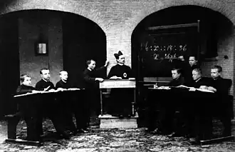 Henri staat voor het schoolbord, rechts. Foto circa 1886