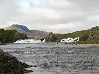 De Laxfoss. Achter het linker deel van de waterval zijn de bebouwing van Bifröst en de Grábrók herkenbaar, achter het rechter deel ligt Baula deels in de wolken verscholen.