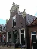 Woonhuis onder zadeldak tegen klokgevel met gesneden deur en bovenlicht, zwierige aanzetkrullen en groot kuifstuk in de plaatselijke Friese trant van de 18e eeuw
