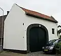 Poortgebouw van mergel, op trapeziumvormige grondslag, afgedekt met een zadeldak. Ellipsboogpoort in baksteen.