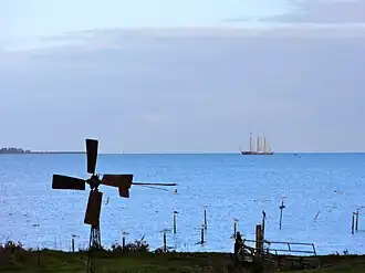 Gezicht op het water met oo de voorgrond een klein windmolentje en op de achtergrons een zeilende driemaster