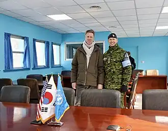 Een foto van Martin Bosma met naast hem de Amerikaanse militair Derek Macaulay. De foto is binnen genomen. Voor hen staat een tafel met daarom drie vlaggen: de Amerikaanse, Zuid-Koreaanse en VN vlag.