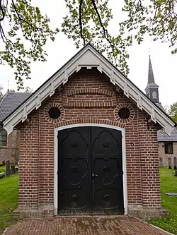 Baarhuis en toegangshek op negentiende-eeuwse kerkhof, rondom de van oorsprong veertiende-eeuwse kerk (RM nr. 39905), die tegenwoordig wordt gebruikt door de Hervormde Gemeente[5]