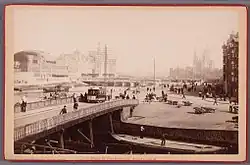 De trambrug van Pieter Oosterhuis met op de achtergrond het Centraal Station in aanbouw; op de brug een Treubpaal van de gemeentetelefoon
