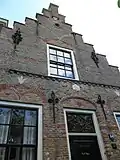 Huis met trapgevel. Sierankers. Gele baksteen met donkerrode lagen en bogen. Muizetand, toppilaster. Benedenstuk gepleisterd