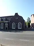 Verdiepingloos pand onder zadeldak; daterend uit de bouwtijd van de kerk, en in een verband daarmee aangelegd