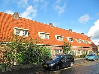 Midden-westelijke woningreeks. Symmetrisch op U-vormige grondslag opgezette woningreeks bestaande uit acht telkens gespiegelde woningen tussen puntgevels onder langgerekt dwars zadeldak