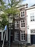 Huis met eenvoudige gevel met rechte kroonlijst met tympaan. Gebeeldhouwde deur. Levensboom in bovenlicht