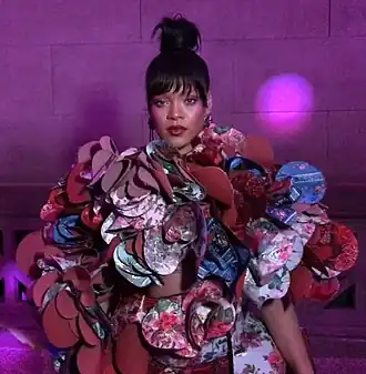 Een ingezoomde foto van Rihanna op het Met Gala van 2017. Ze is gekleed in een kleurrijke, driedimensionale jurk ontworpen door Rei Kawakubo voor het modemerk Comme des Garçons. De jurk bestaat uit een reeks overlappende, bloemvormige panelen van stof in felle kleuren en drukke patronen, waaronder rood, roze, blauw en paars. De stofpanelen creëren een driedimensionaal effect. Rihanna draagt haar haar in een knot en haar oogschaduw, lipstick en blush zijn verschillende paarstinten. De foto is gekropt en toont alleen haar bovenlichaam. De achtergrond is paars.