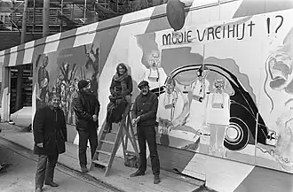 Ria Rettich op een trapje bij schilderijen op een schutting om de Nieuwe Kerk voor de tentoonstelling "Mooie Vrijheid? 19 maart 1970