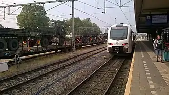 Arriva FLIRT op het station. Links passeert een goederentrein met legervoertuigen het station