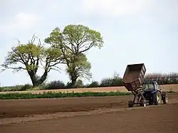 Landbouwmachine met kipper op akker met 2 bomen op de achtergrond