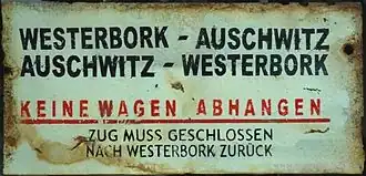 Een licht gekleurd koersbord "met in zwarte hoofdletters het opschrift "Westerbork - Auschwitz, Auschwitz - Westerbork", rood onderstreept in het Duits "Keine Wagen abhangen" en in kleinere donkere hoofdletters daaronder "Zug muss geschlossen nach Westerbork zurück"