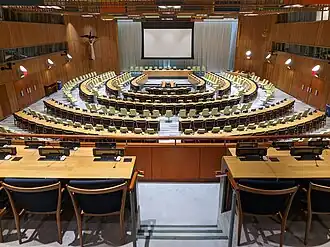 Overzichtsfoto van de vergaderzaal van de Trustschapsraad van de Verenigde Naties. De stoelen op de foto zijn geplaatst in de vorm van een halve cirkel. De wanden van de zaal zijn bekleed met hout. Aan het plafond hangt een houten constructie met gekleurde kubussen waar lampen in zijn verwerkt.
