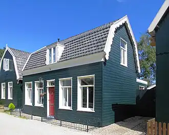 Dwars geplaatst houten huis met gerabatte wanden met zolderverdieping onder mansarde kap; kroonlijst langs voorgevel; T-vensters