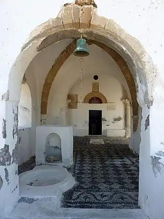 De foto toont een witte boog waardoor men onder een bel door naar het portiek van de kerk en de deur naar de hoofdruimte kan kijken. De vloer van de veranda bestaat uit een tweekleurig mozaïek.